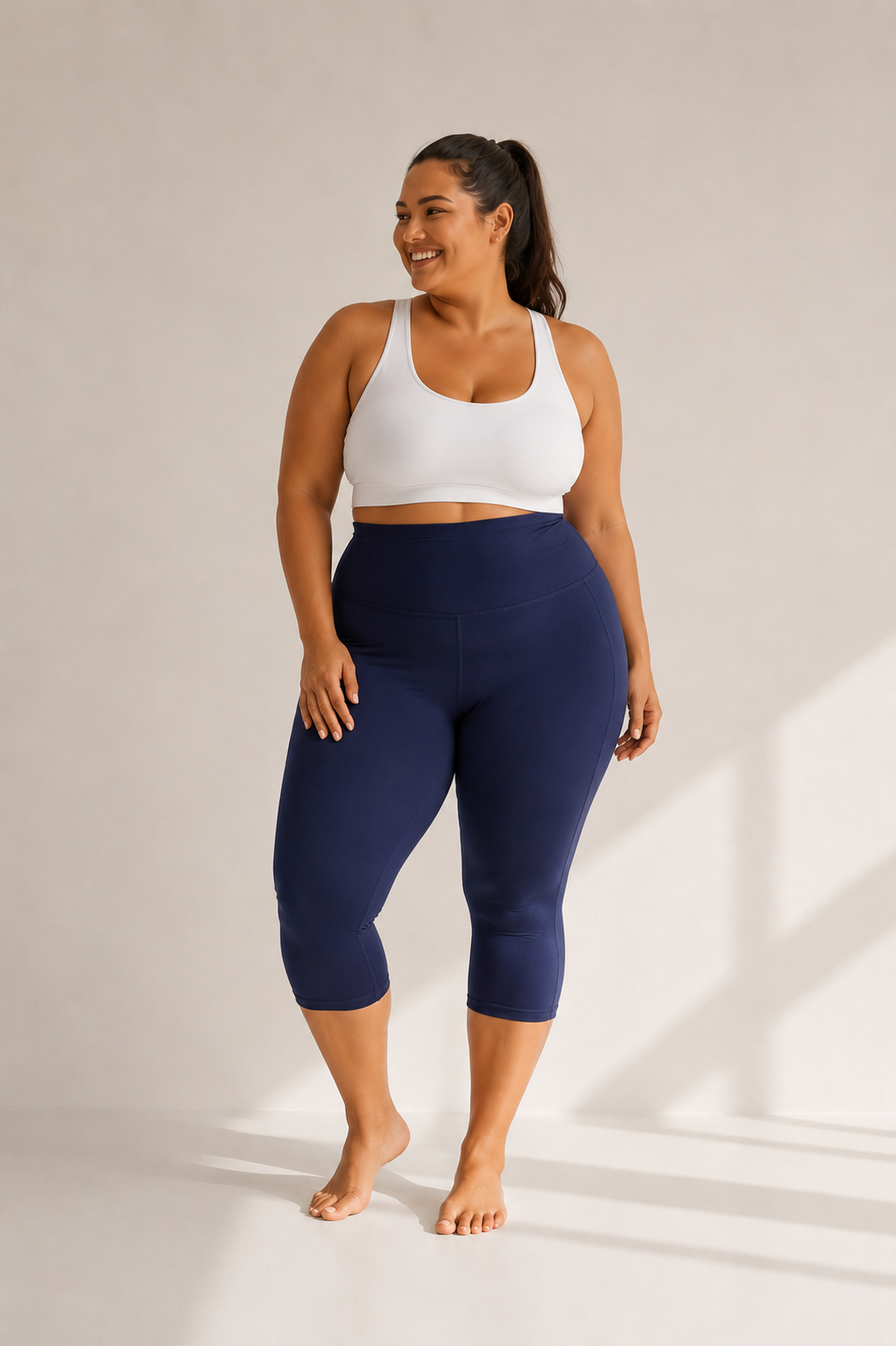 Woman wearing Deep Ocean Blue high waisted pocket capri leggings by Barefoot & Busy with a white sports bra in a studio setting